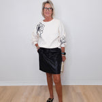 Woman wearing a white sweater with black floral designs and a black skirt against a white wall.