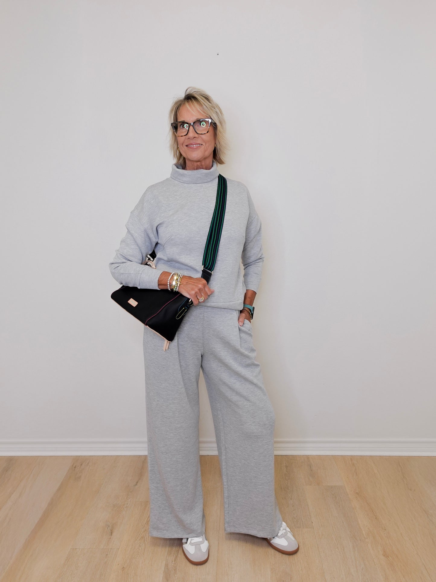 Woman wearing a gray outfit with a black handbag against a white wall.
