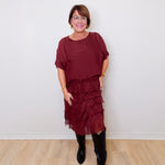 Woman wearing a burgundy dress with ruffled layers and black boots against a white wall.