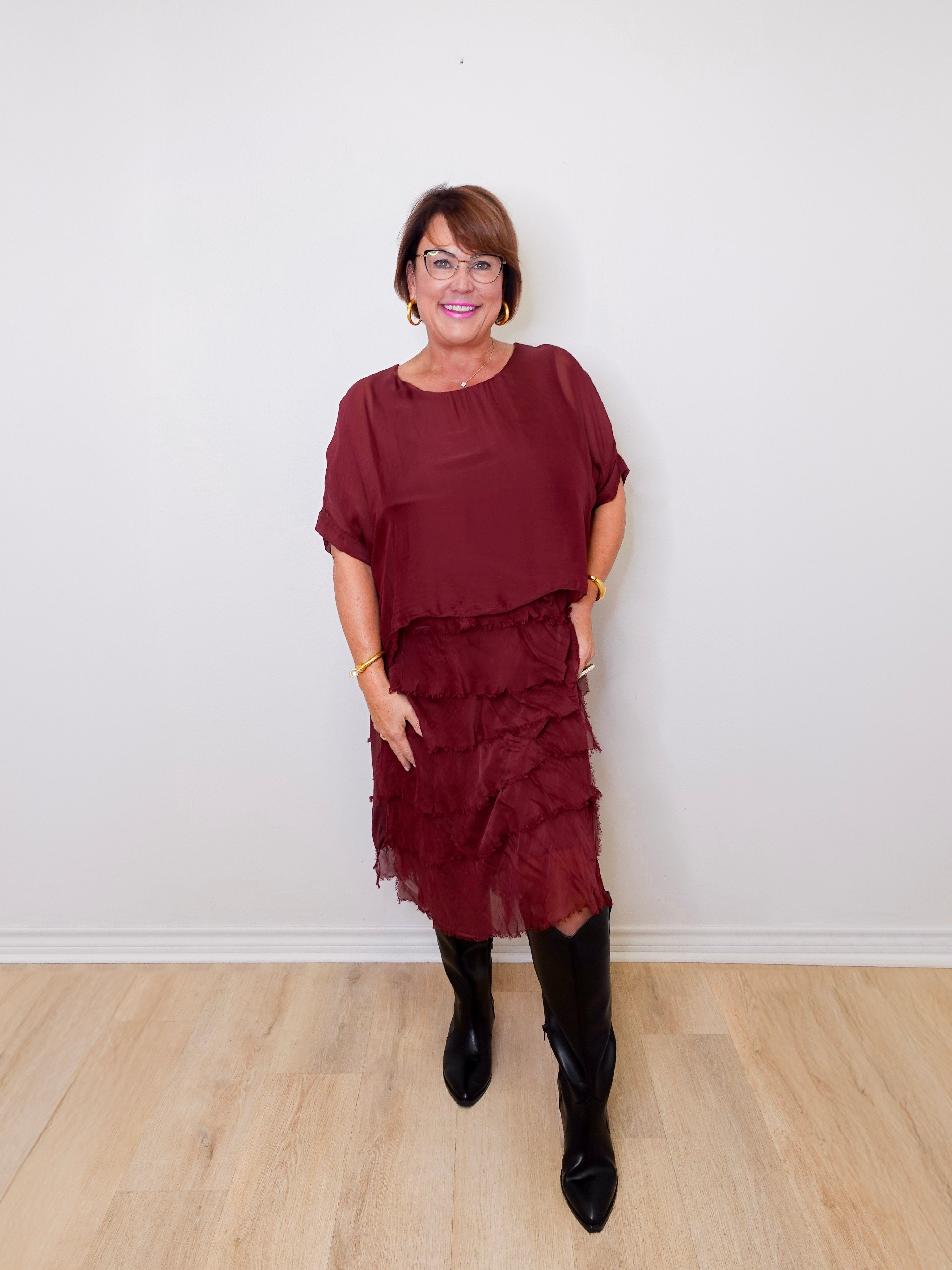 Woman wearing a burgundy dress with ruffled layers and black boots against a white wall.