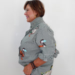 A woman shown in profile wearing a black and white gingham button-up shirt with colorful duck illustrations and light-wash jeans, standing against a plain white wall.