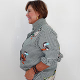 A woman shown in profile wearing a black and white gingham button-up shirt with colorful duck illustrations and light-wash jeans, standing against a plain white wall.