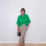 Woman wearing a green shirt and beige pants against a white wall.