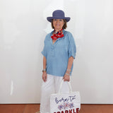 Woman wearing a blue shirt, white pants, and a purple hat, holding a tote bag with text.