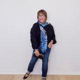Woman wearing a black jacket, blue jeans, and a patterned scarf against a white wall.