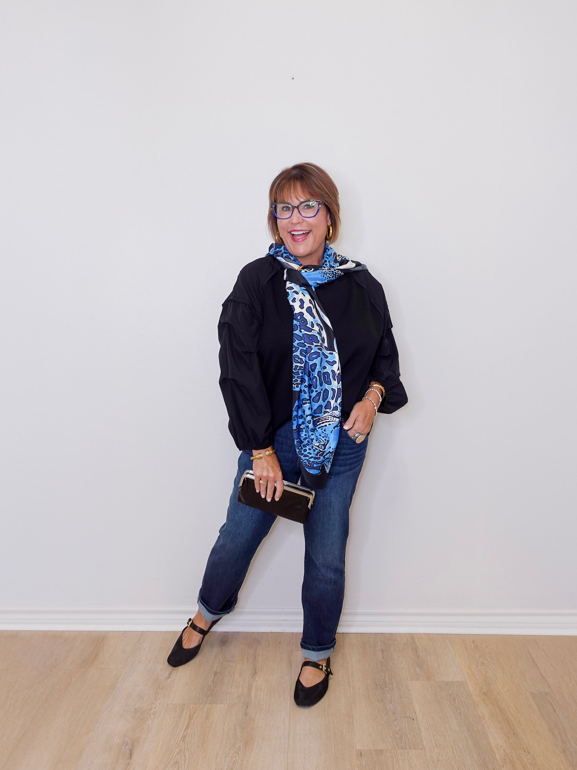 Woman wearing a black jacket, blue jeans, and a patterned scarf against a white wall.