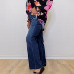Person wearing a floral top and blue jeans standing against a white wall.