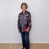 Woman wearing a patterned jacket and jeans standing against a white wall.