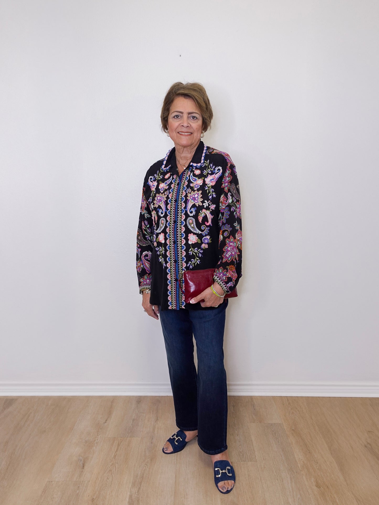 Woman wearing a patterned jacket and jeans standing against a white wall.