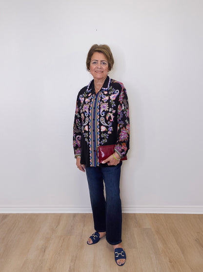 Woman wearing a patterned jacket and jeans standing against a white wall.