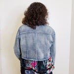 Person wearing a denim jacket with floral patchwork design on a plain background