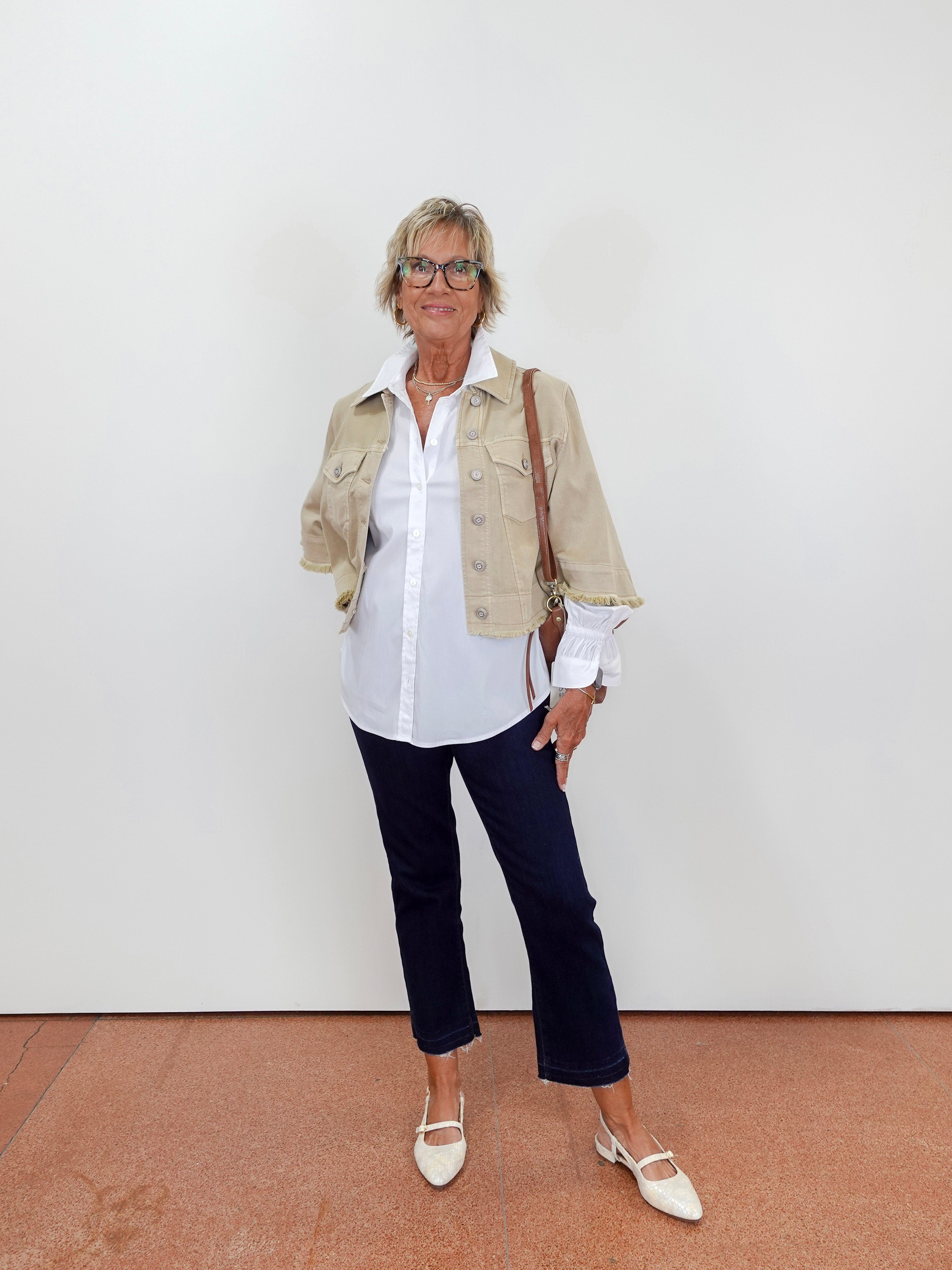Woman wearing a beige jacket, white shirt, dark pants, and white shoes against a plain background