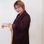 Woman wearing a brown blazer and glasses, standing against a white wall.