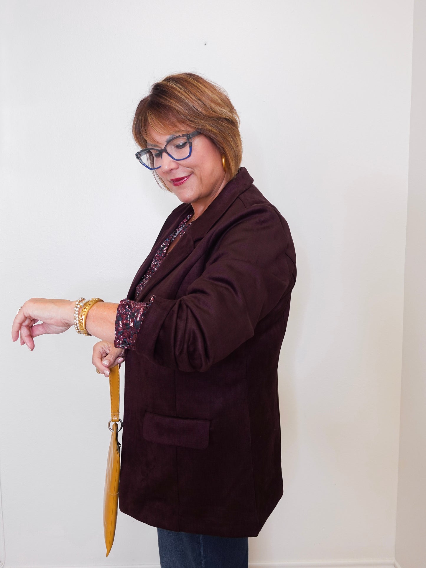 Woman wearing a brown blazer and glasses, standing against a white wall.