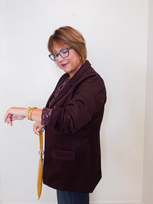 Woman wearing a brown blazer and glasses, standing against a white wall.