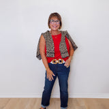 Woman wearing a red top, leopard print vest, blue jeans, and black shoes against a white wall.
