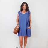 Woman wearing a blue dress with ruffled sleeves against a white wall.