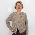 An older woman with short blonde hair stands against a plain white wall wearing a textured tweed long-sleeve top in beige, brown, and cream tones, paired with dark brown trousers.