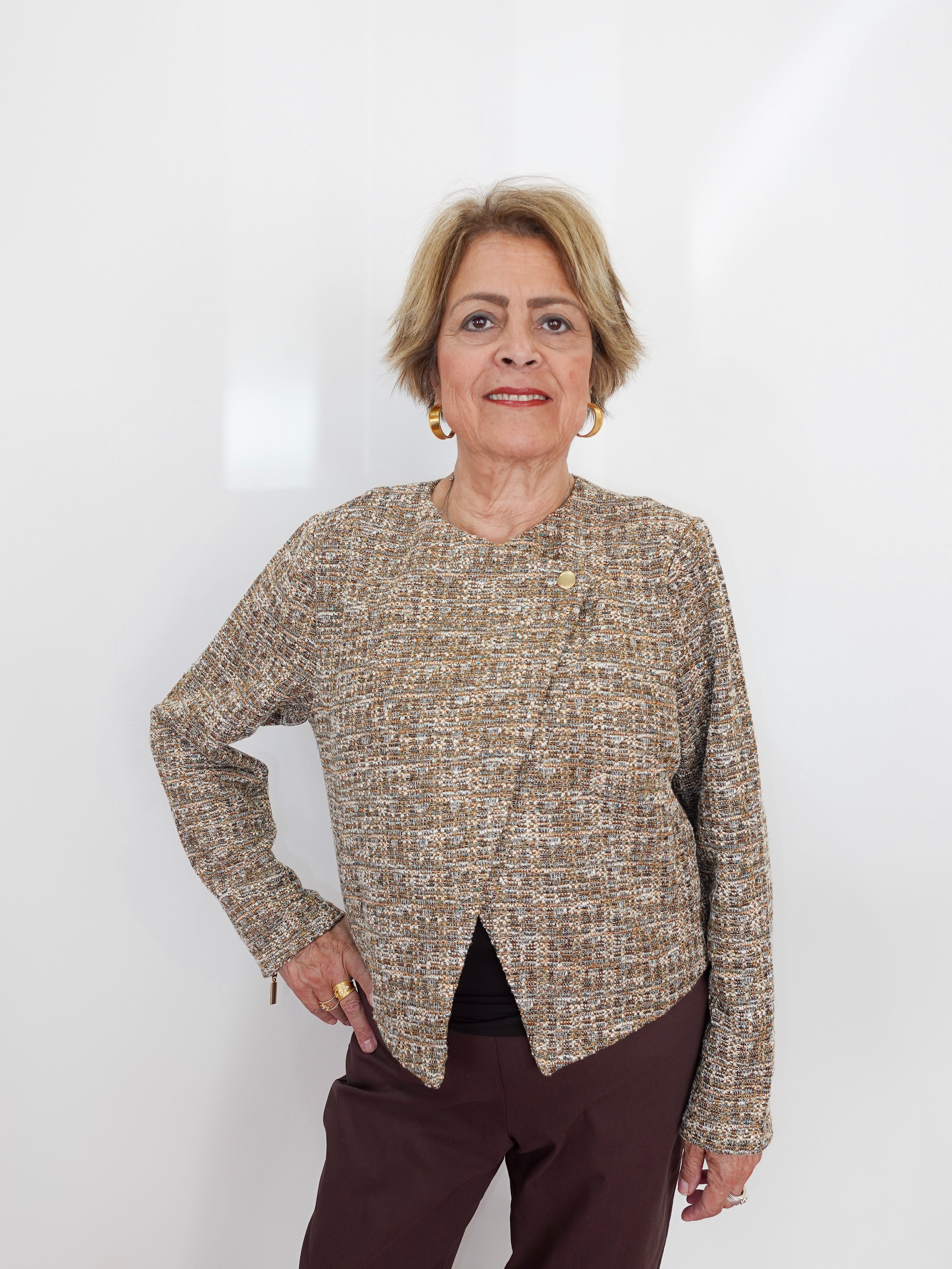 An older woman with short blonde hair stands against a plain white wall wearing a textured tweed long-sleeve top in beige, brown, and cream tones, paired with dark brown trousers.
