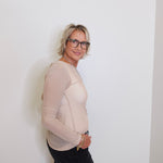 Person wearing a beige long-sleeve top and black pants against a white wall