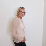 Person wearing a beige long-sleeve top and black pants against a white wall