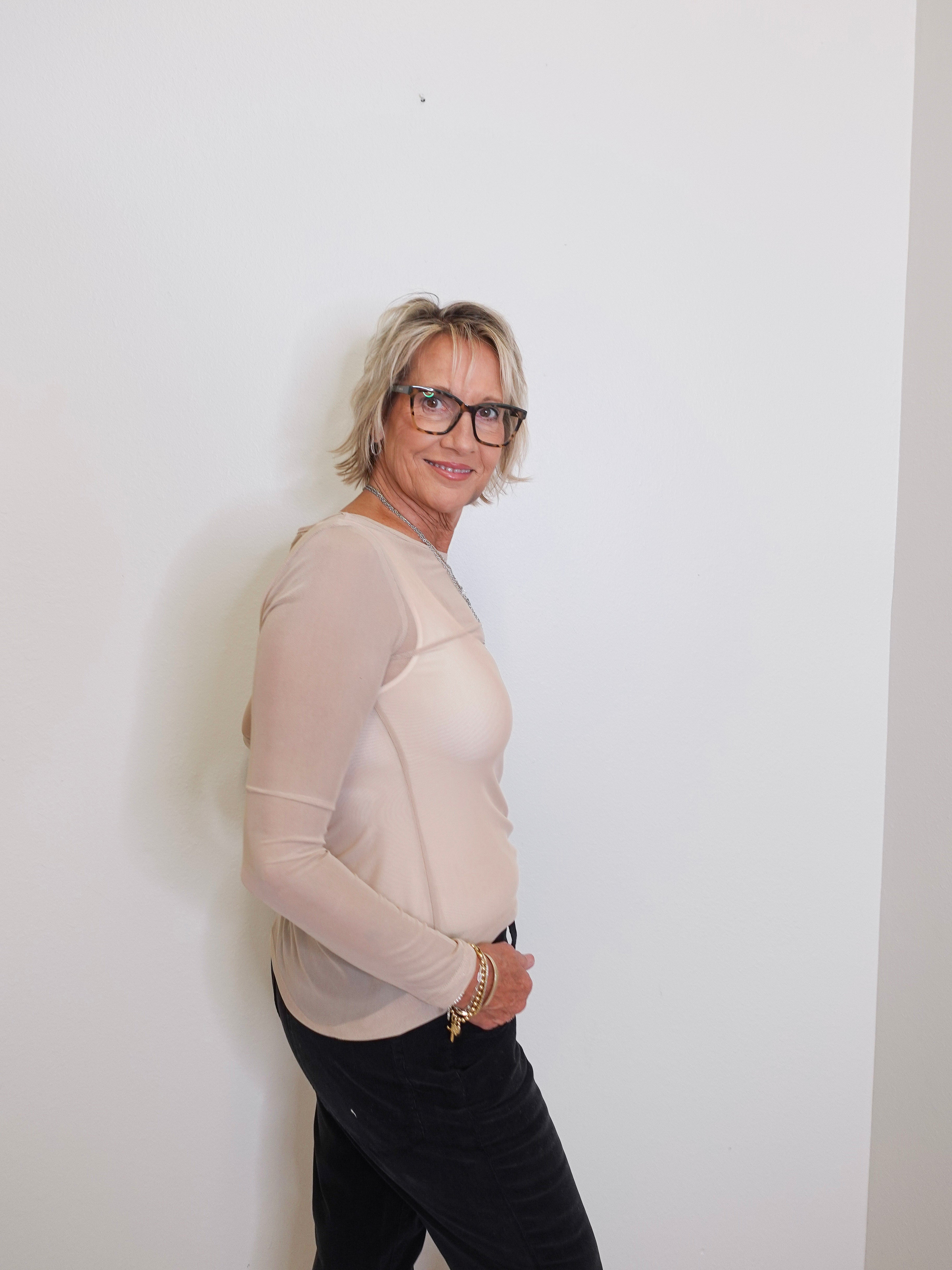 Person wearing a beige long-sleeve top and black pants against a white wall