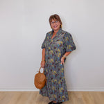 Woman wearing a floral dress holding a brown handbag against a white wall.