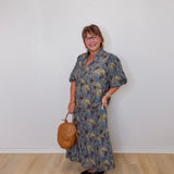 Woman wearing a floral dress holding a brown handbag against a white wall.
