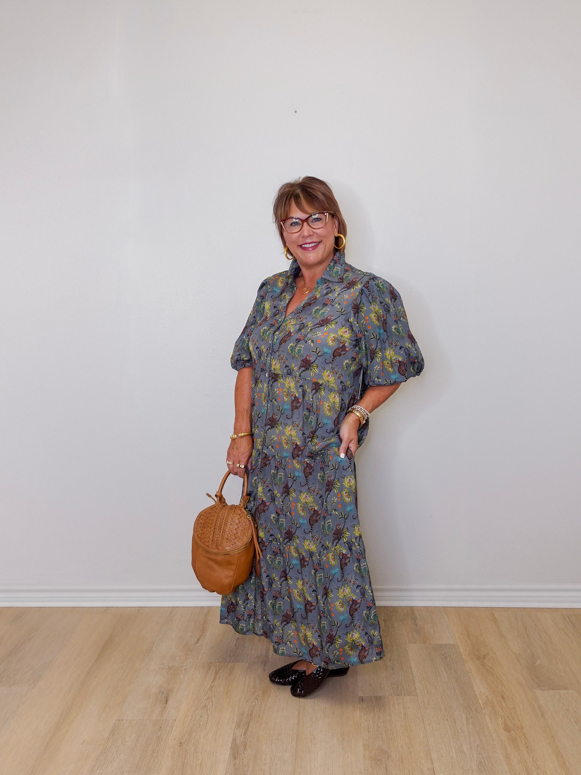 Woman wearing a floral dress holding a brown handbag against a white wall.