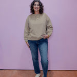 Woman wearing a beige sweater and blue jeans standing against a pink wall.