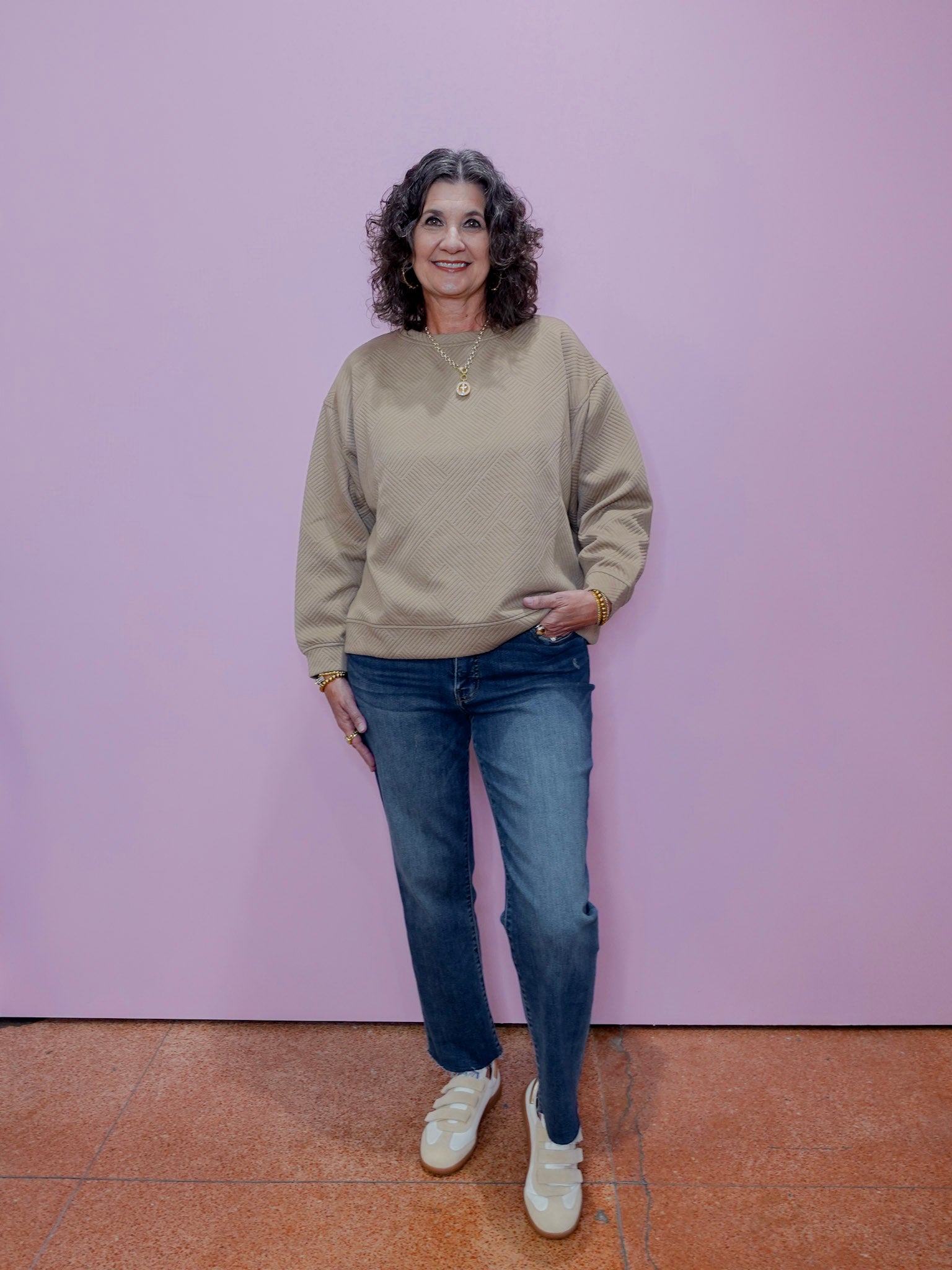 Woman wearing a beige sweater and blue jeans standing against a pink wall.