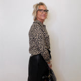 Person wearing a leopard print shirt and black skirt holding a black bag against a white wall.