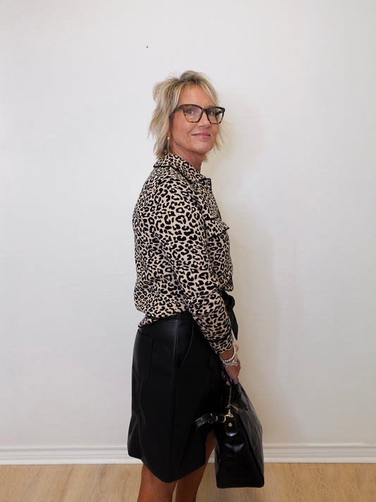 Person wearing a leopard print shirt and black skirt holding a black bag against a white wall.