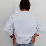 A person viewed from behind wearing a light gray heather crewneck sweatshirt and light blue jeans, standing against a plain white wall.