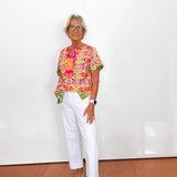 Woman wearing a colorful top and white pants standing against a white wall.