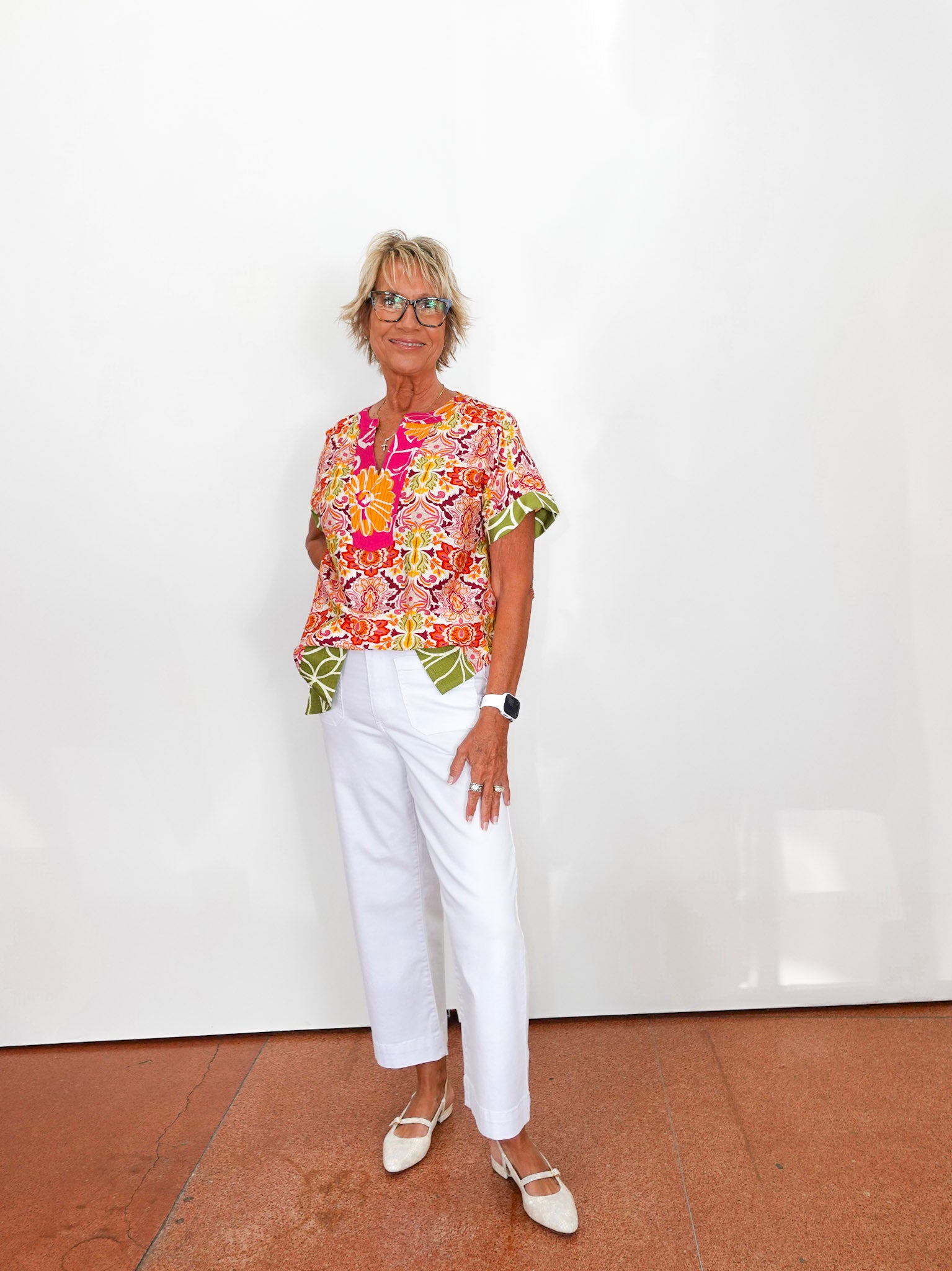 Woman wearing a colorful top and white pants standing against a white wall.