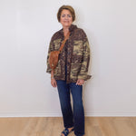 Woman wearing a camouflage jacket and blue jeans standing against a white wall.