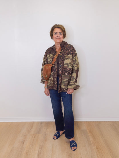 Woman wearing a camouflage jacket and blue jeans standing against a white wall.