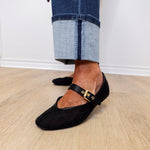 Person wearing black flat shoes with gold buckles and rolled-up jeans on a wooden floor.