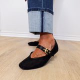 Person wearing black flat shoes with gold buckles and rolled-up jeans on a wooden floor.