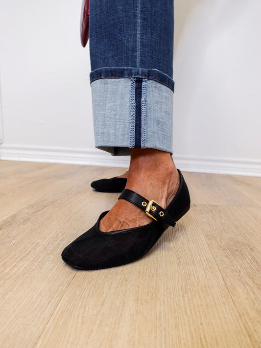Person wearing black flat shoes with gold buckles and rolled-up jeans on a wooden floor.