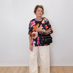 Woman wearing a floral top and white pants standing against a white wall.
