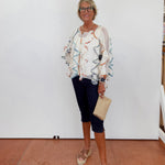 Woman wearing a patterned blouse and dark pants, standing against a white wall.