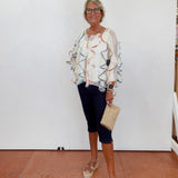 Woman wearing a patterned blouse and dark pants, standing against a white wall.