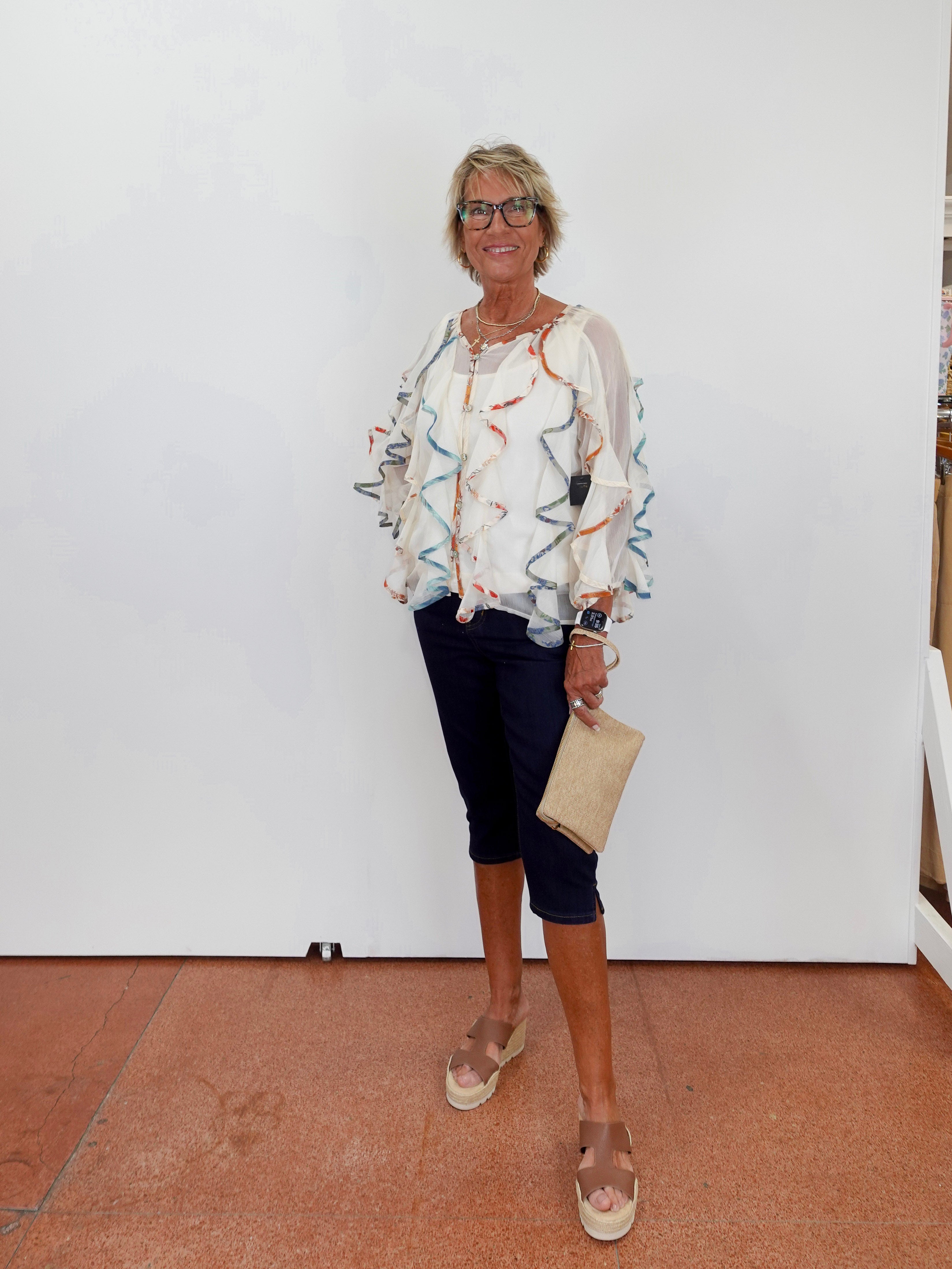 Woman wearing a patterned blouse and dark pants, standing against a white wall.