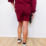 Person wearing a maroon outfit with shorts and a top, standing on a wooden floor against a white wall.