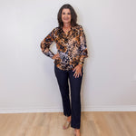 Woman wearing a leopard print blouse and navy pants standing against a white wall.