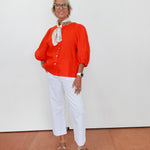 Woman wearing an orange blouse, white pants, and brown shoes on a plain background