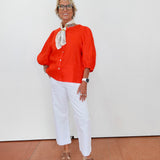 Woman wearing an orange blouse, white pants, and brown shoes on a plain background