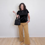 Woman wearing a black top and beige pants holding a black handbag against a white wall.